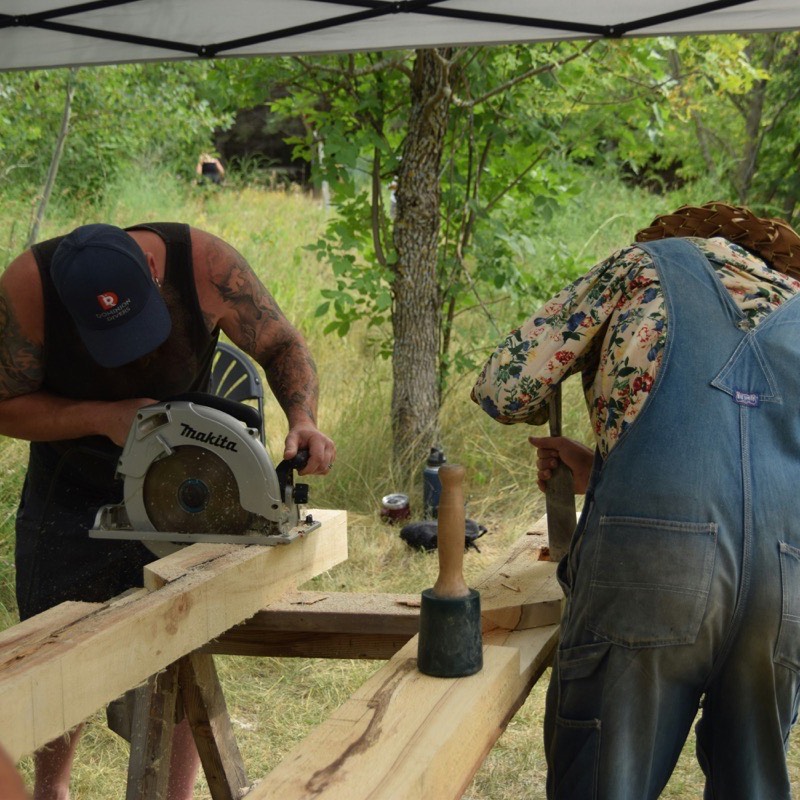 Zwei Personen arbeiten an einem Holzbalken mit Kreissäge und Meißel